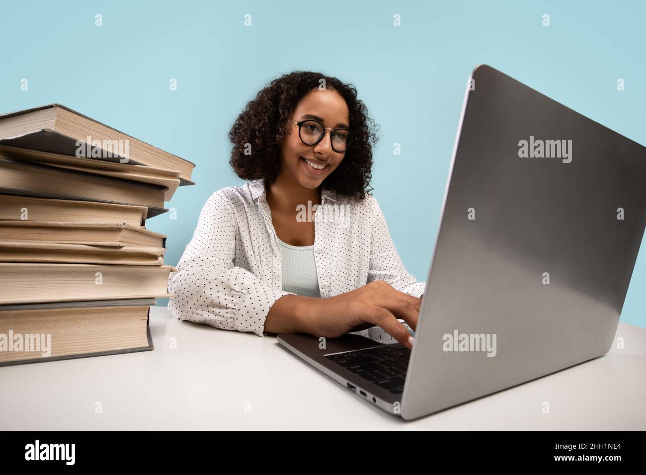 Positive young black female student with heap of books working on ...