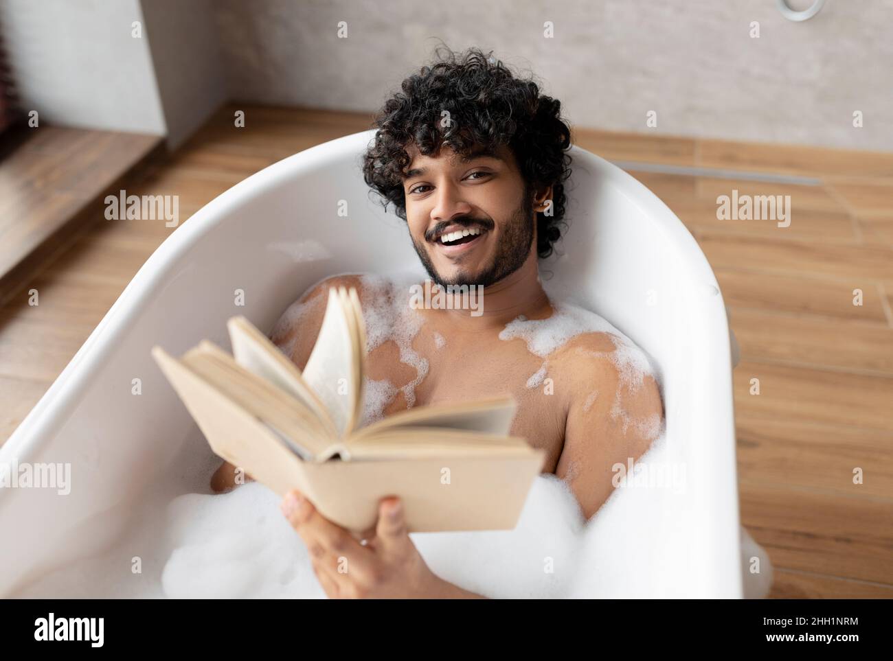 Happy indian man lying in foamy bath and reading book in relaxing
