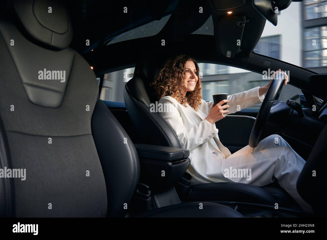 Cheerful Caucasian business woman have breakfast during time for driving corporate automobile, happy female entrepreneur with takeaway coffee to go smiling while steering vehicle transport Stock Photo