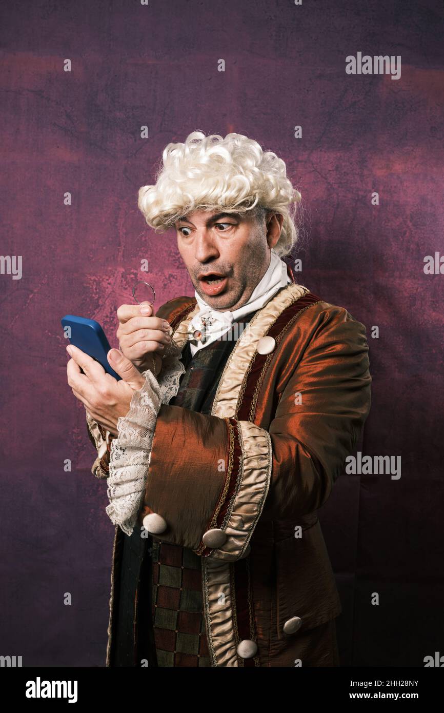 Man in classical renaissance clothes with a surprised expression on his face while looking at the mobile phone screen with a monocle. Stock Photo