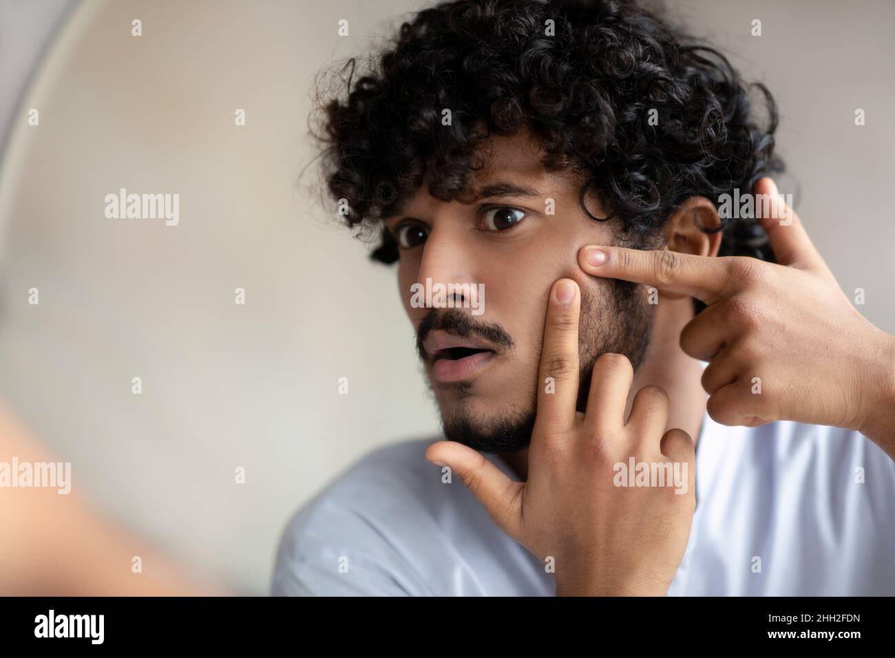 Portrait of handsome indian man touching his face, pressing pimple on face with fingers, looking at mirror, closeup Stock Photo