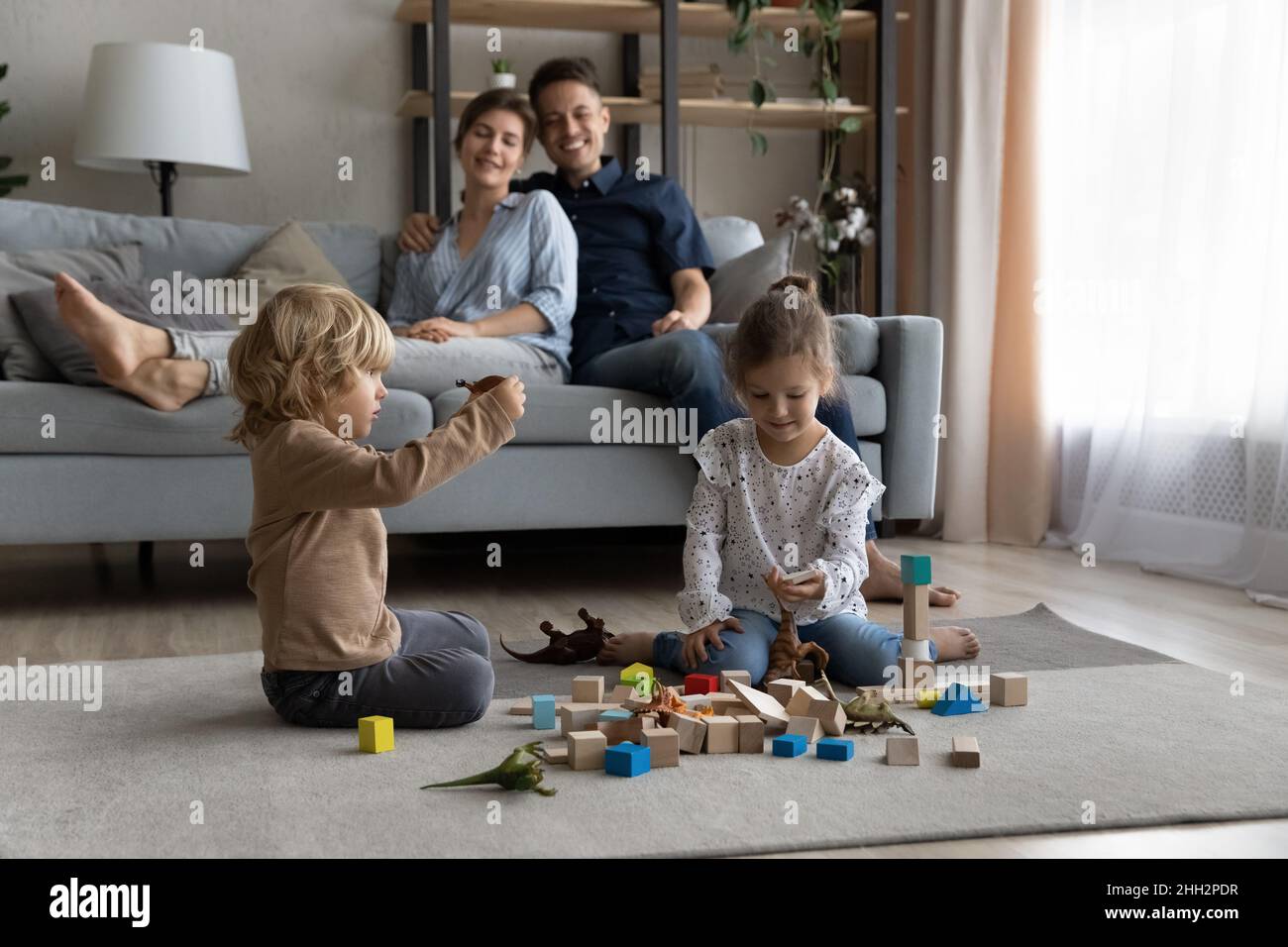 Happy little kids playing toys while parents resting on sofa. Stock Photo