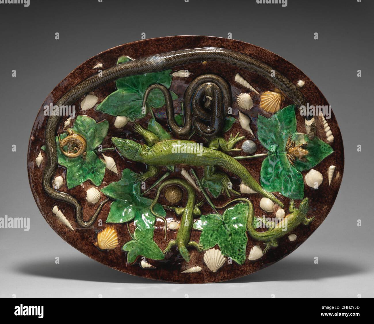 Oval basin with lizards, shells, and ivy leaves with brown background ca. 1865–1875 Georges Pull Georges Pull created some of the most sophisticated examples of Palissy ware, ceramics made in the nineteenth century inspired by the work of the Renaissance potter Bernard Palissy (1510-c.1589). The reverse of this dish shows deep impressions left by the lizard and the snake, indicating Pull, like Palissy, created the basin and the creatures decorating its surface from a single mold.. Oval basin with lizards, shells, and ivy leaves with brown background. French, Paris. ca. 1865–1875. Glazed earthe Stock Photo