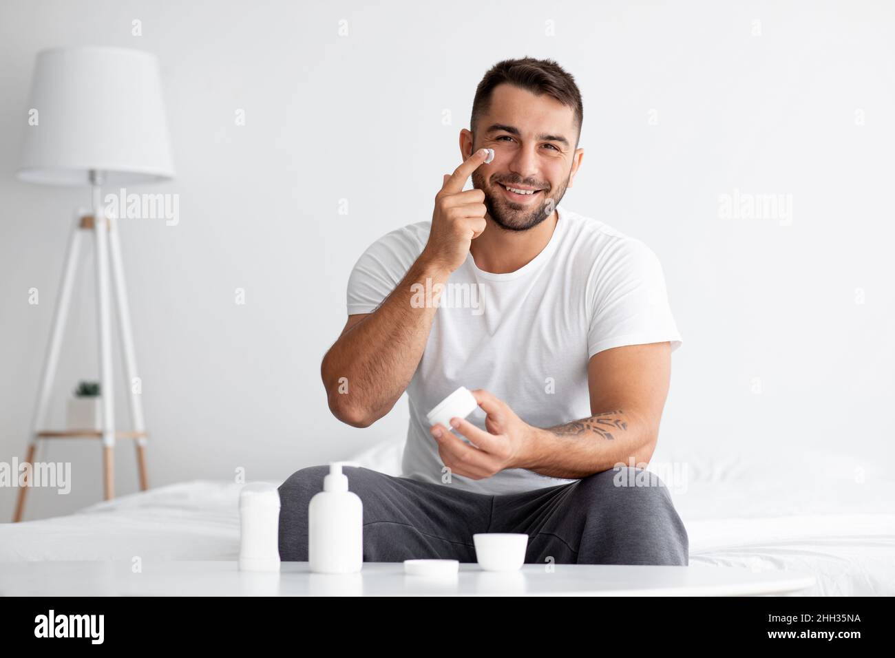Cheerful young attractive guy with beard applies cream on his face in minimalist bedroom interior with jars at table Stock Photo