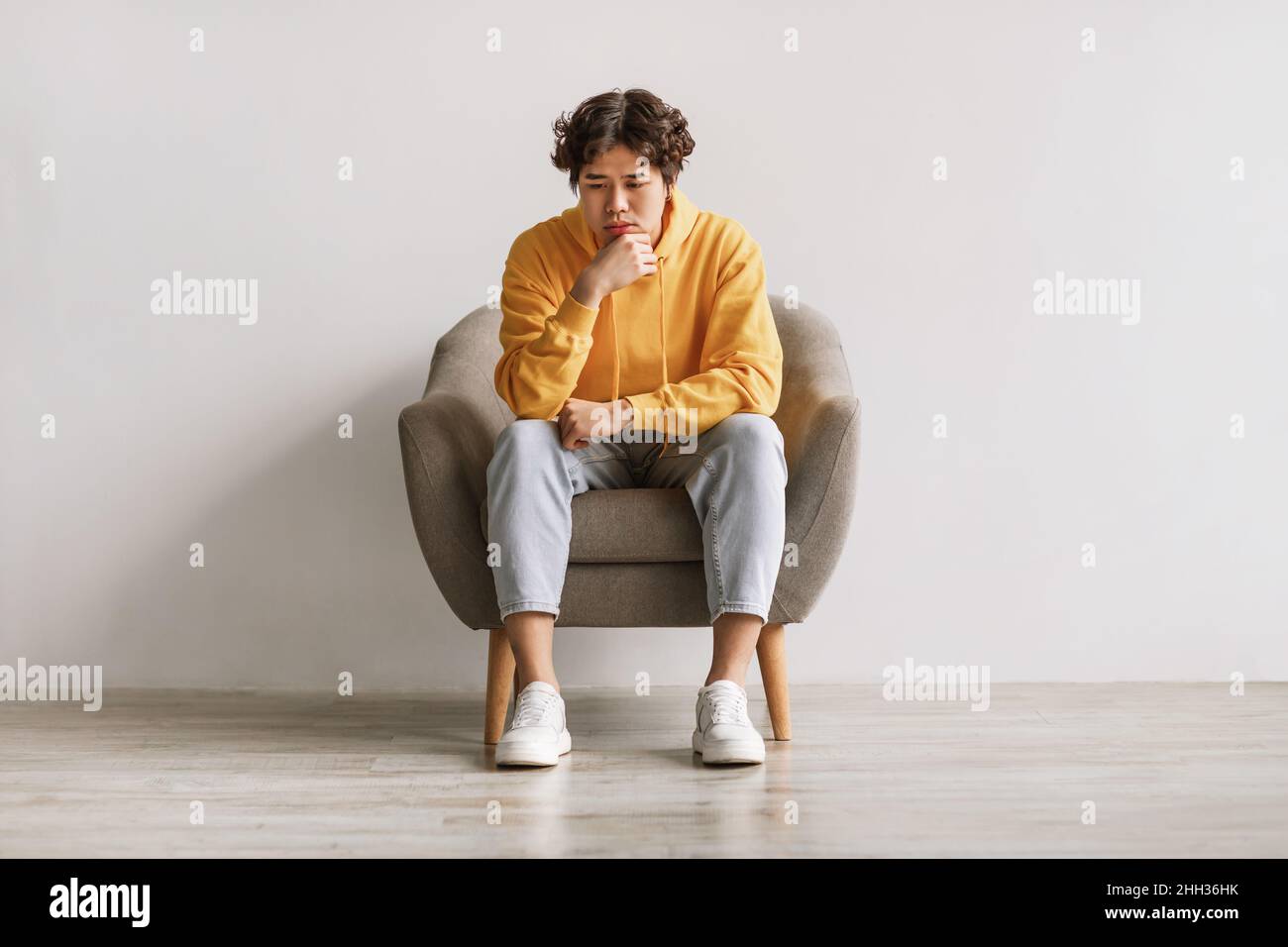 Thoughtful Asian man sitting in armchair, feeling sad or tired, having problem, suffering from depression or stress Stock Photo