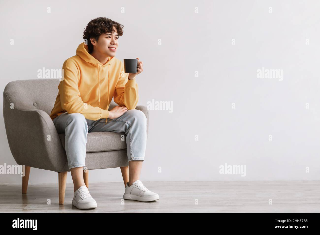 Handsome young Asian man drinking coffee, having break, chilling in armchair with hot drink against white studio wall Stock Photo