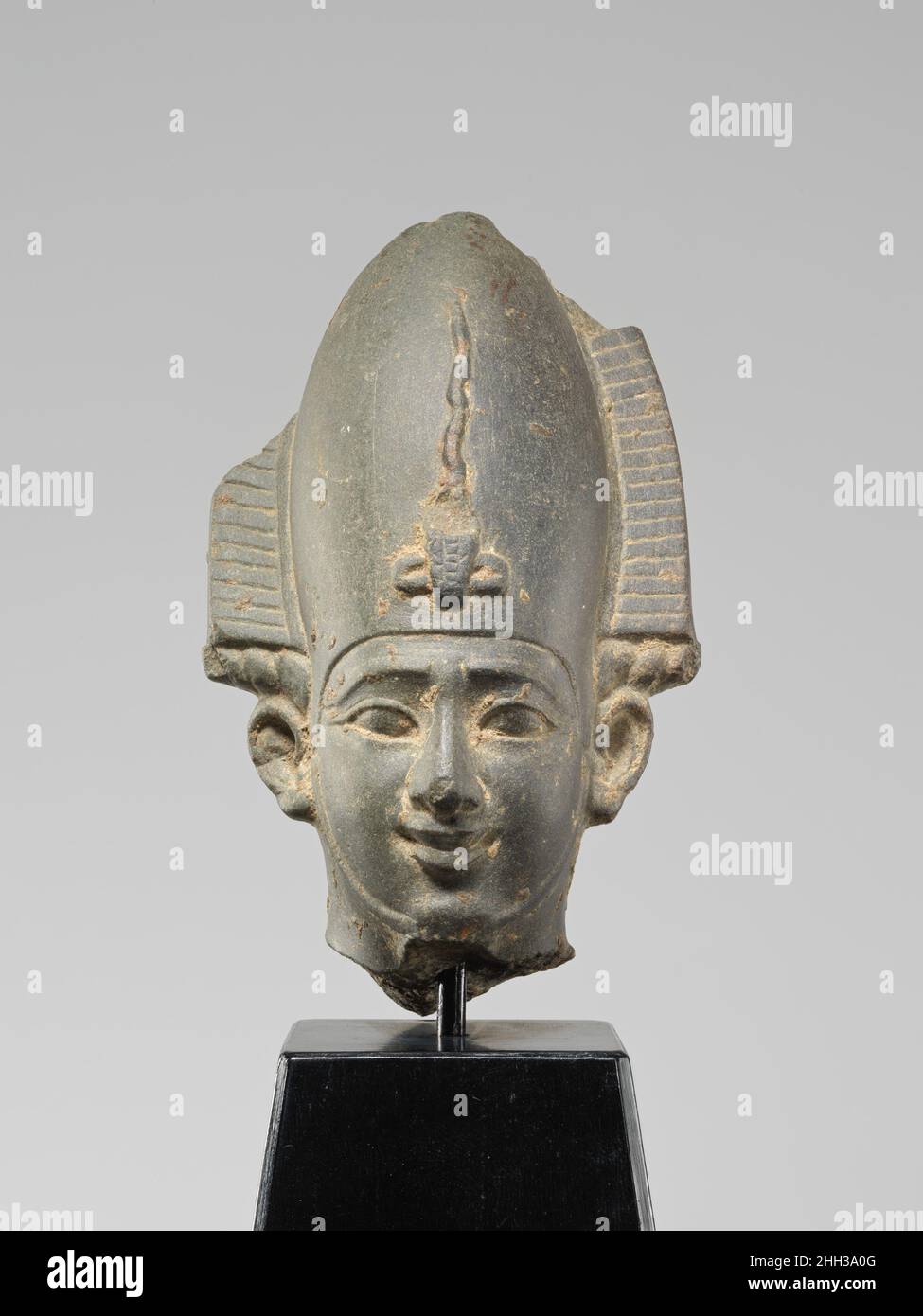 Head from a figure of Osiris ca. 664–525 B.C. Late Period (Saite) This head is from a statuette of Osiris, principal god of the afterlife and ruler of the Netherworld. The deity is shown wearing his typical "atef" crown, a tall crown flanked by ram's horns and ostrich plumes and protected by a hooding cobra. His face is placid and his serene affect enhanced by a slight smile. Only the very top of his false beard and the straps that would have held it in place are preserved. The tip of the crown, as well as the tops of the feathers and the tips of the horns that flanked it are gone, and the hea Stock Photo