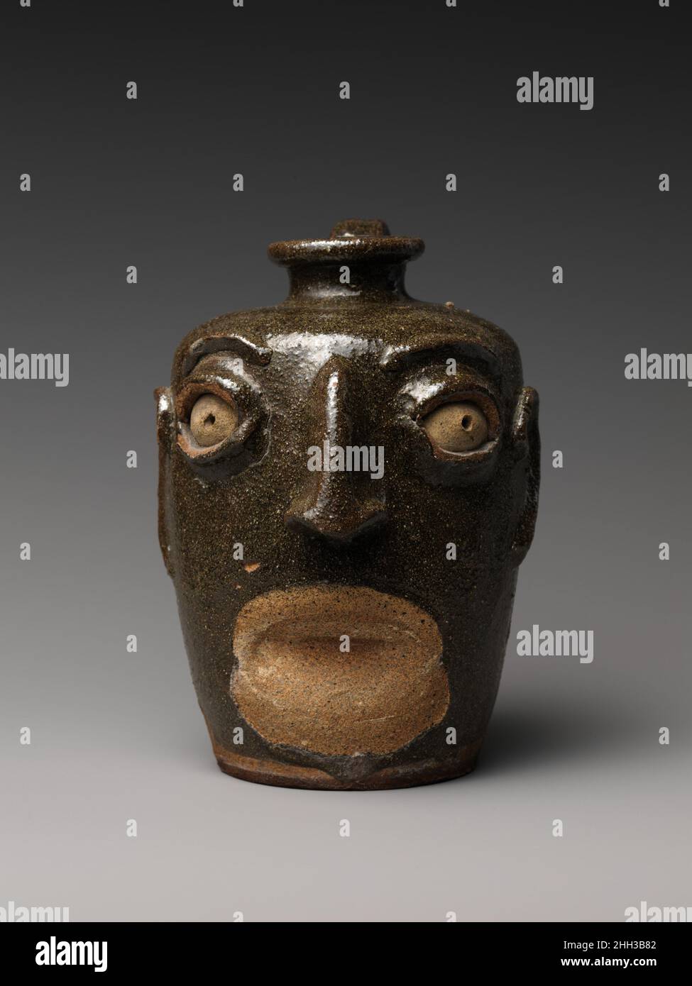 Face Jug ca. 1860–80 Unidentified Edgefield District potter Face jugs were made by African American slaves and freedmen working in potteries in the Edgefield District of South Carolina, an area of significant stoneware production in the nineteenth century. The distinctive features of the jugs, notably the kaolin inserts for the eyes, relate in style and material to ritualistic objects of the Congo and Angola region of western Africa, whence many slaves in South Carolina descended. This jug is missing its teeth and lips—a common loss, sometimes occurring as early as the firing stage.. Face Jug. Stock Photo