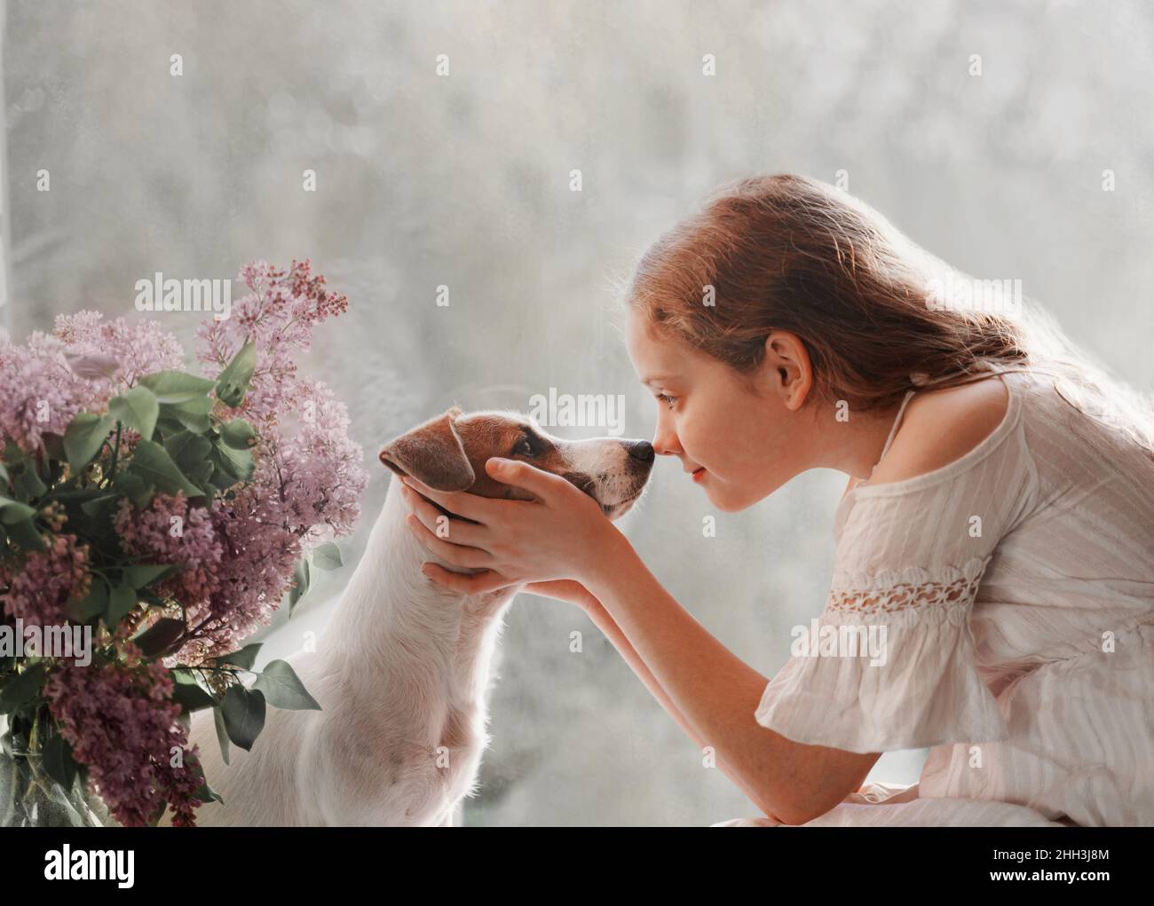 Cute little girl looks into the eyes of a dog. Two friends sitting on the window. Friendship, care, happy childhood concept. Stock Photo