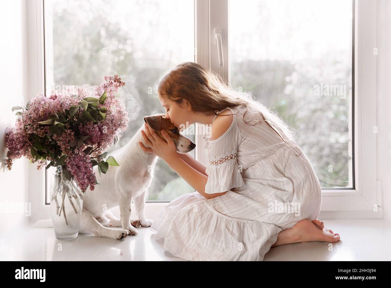 Cute little girl kissing a dog. Two friends sitting on the window. Friendship, care, happy childhood concept. Stock Photo