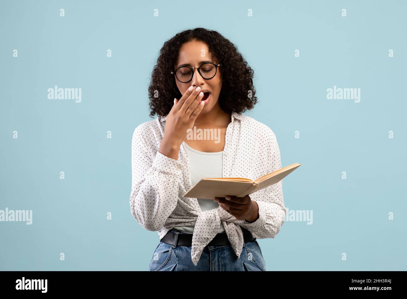 Tired black female student yawning while reading book on blue studio background Stock Photo