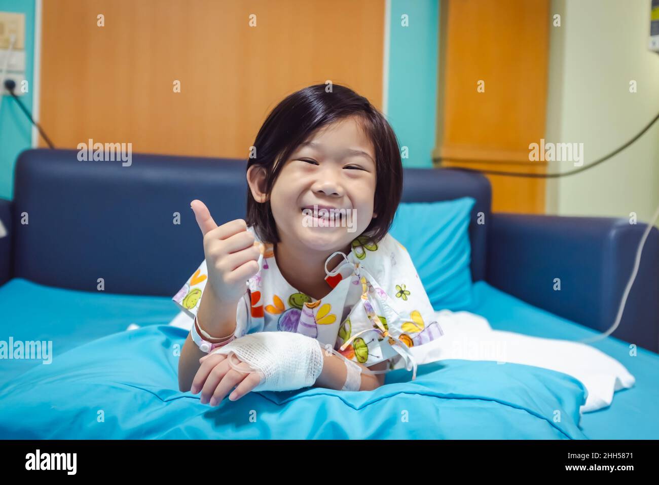 Illness asian child smiling happily and showing thumb up hand sign. Girl admitted in hospital while saline intravenous (IV) on hand. Health care stori Stock Photo
