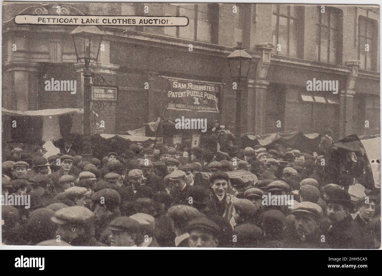 Petticoat Lane clothes auction: large crowd (mostly men in flat caps) in the street, early 20th century. A sign for Sam Rubinstein's patent puzzle purses is in the background Stock Photo