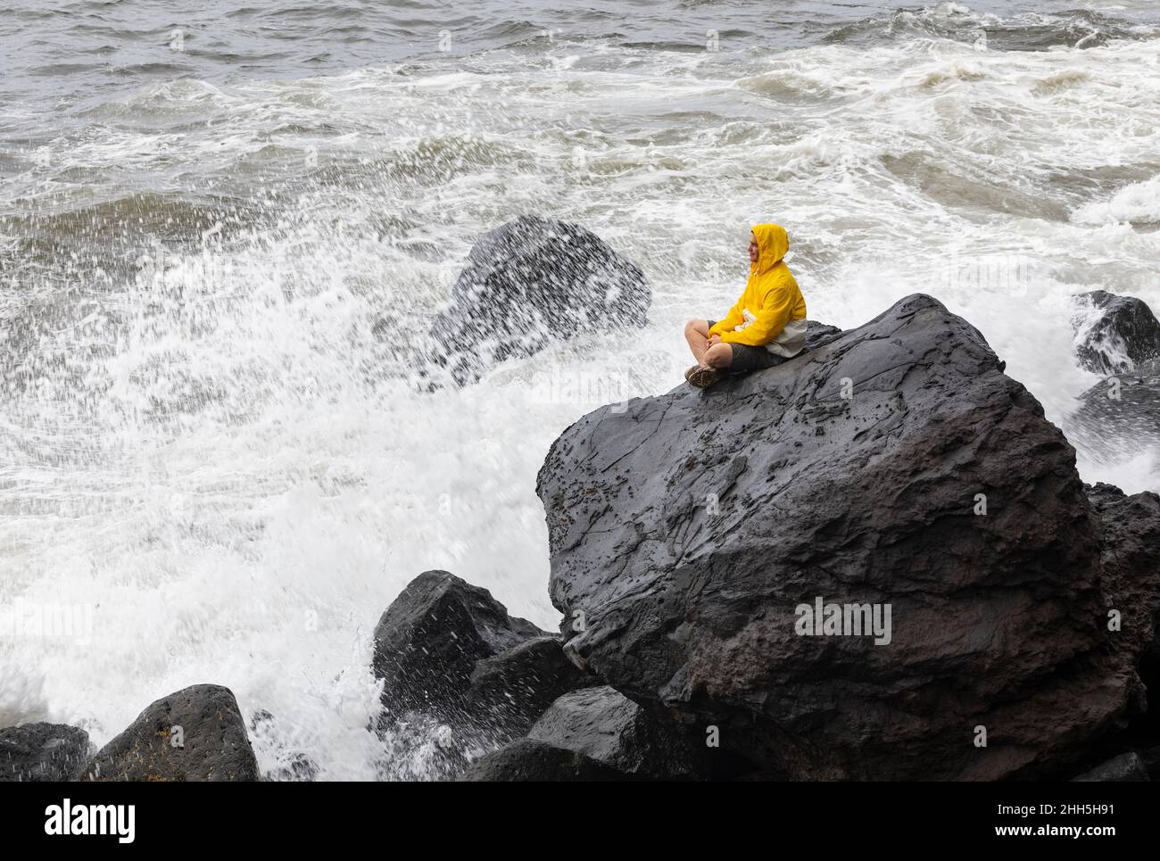 Young man wearing raincoat sitting on rock by waves splashing in sea, Rocha Da Relva, San Miguel Island, Azores, Portugal Stock Photo