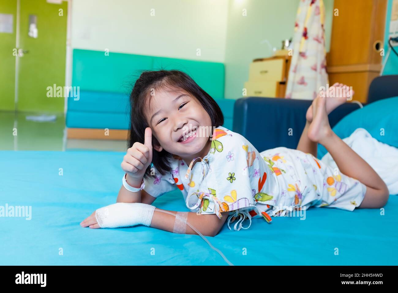 Illness asian child smiling happily and showing thumb up sign. Girl admitted in hospital while saline intravenous (IV) on hand. Health care stories. Stock Photo