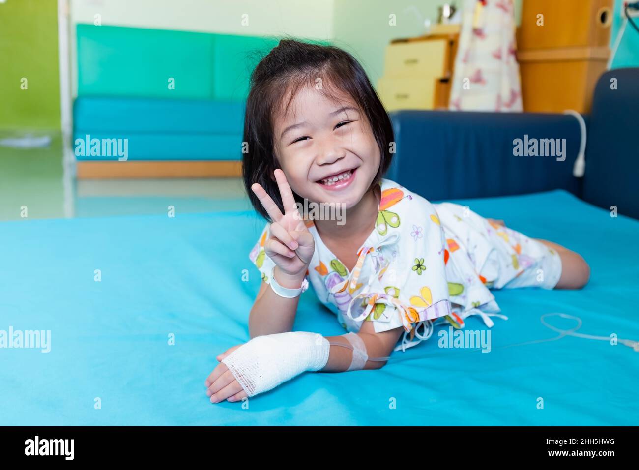 Illness asian child smiling happily and showing v-sign. Girl admitted in hospital while saline intravenous (IV) on hand. Health care stories. Stock Photo