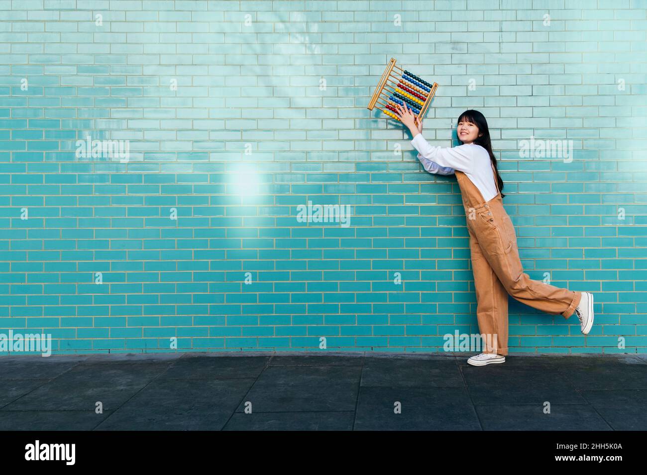 Smiling young woman holding abacus in front of turquoise brick wall Stock Photo