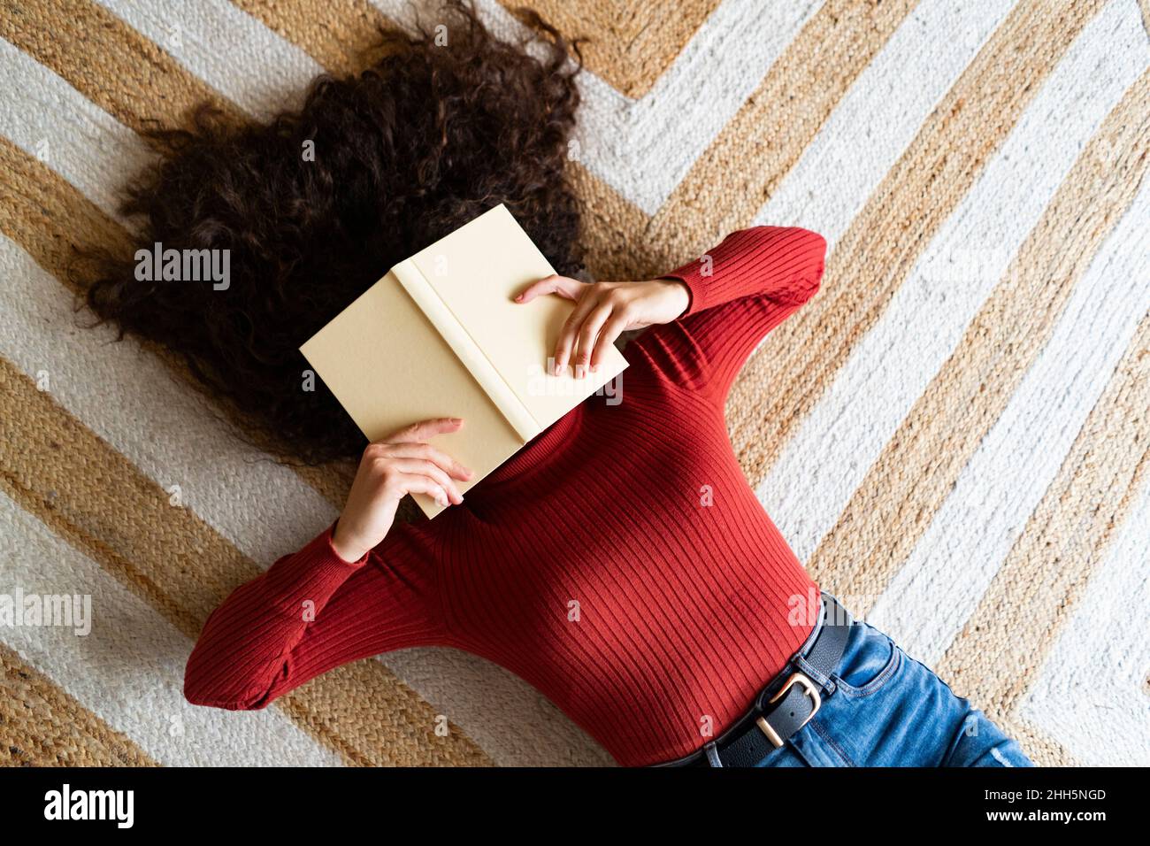 Woman covering face with book lying on carpet at home Stock Photo