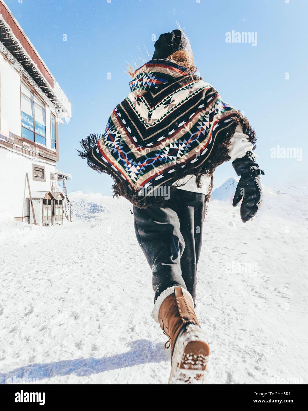 Man wearing poncho running on snow towards mountain hut Stock Photo