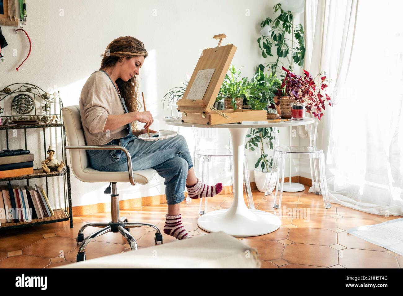 Artist mixing colors in palette sitting on chair in front of easel at home Stock Photo