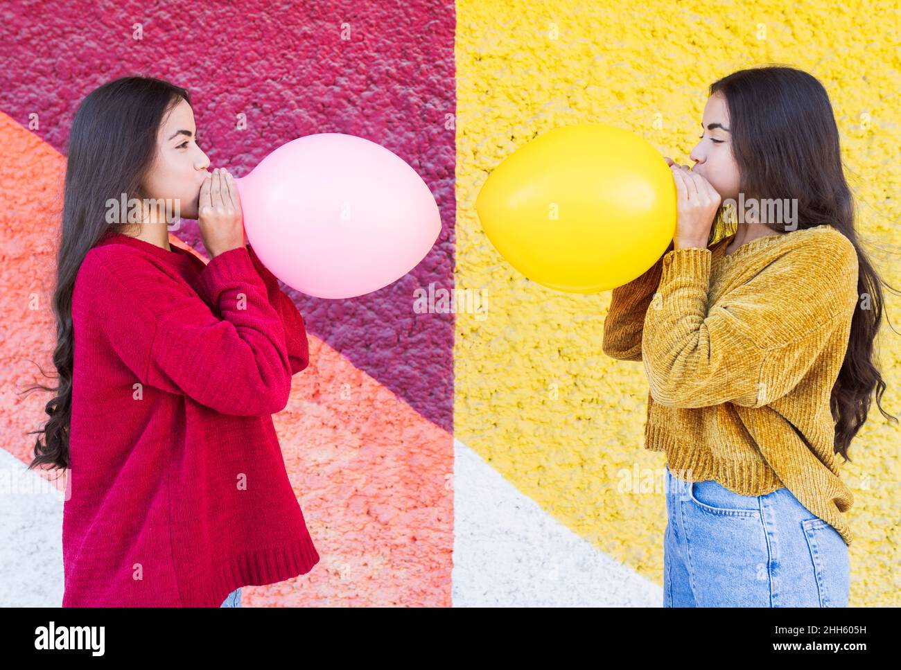 Playful sisters blowing up balloon next to multi colored wall Stock Photo