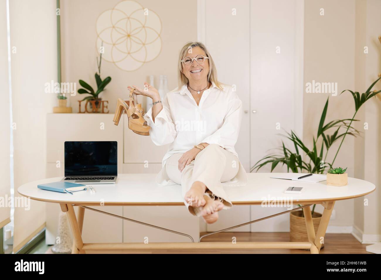 Happy businesswoman holding high heels sitting on desk in office Stock