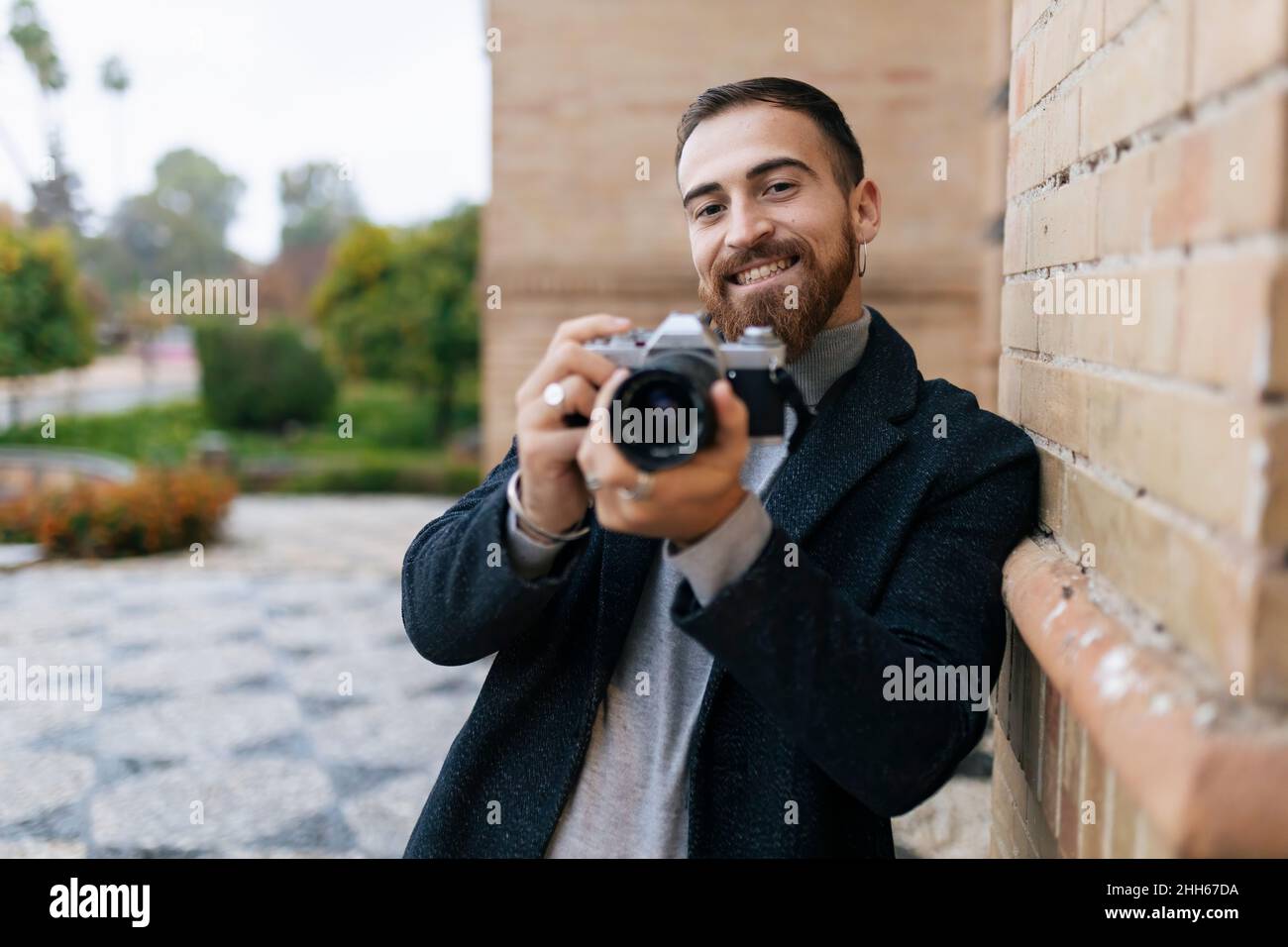 Happy young bearded man with camera leaning on wall Stock Photo