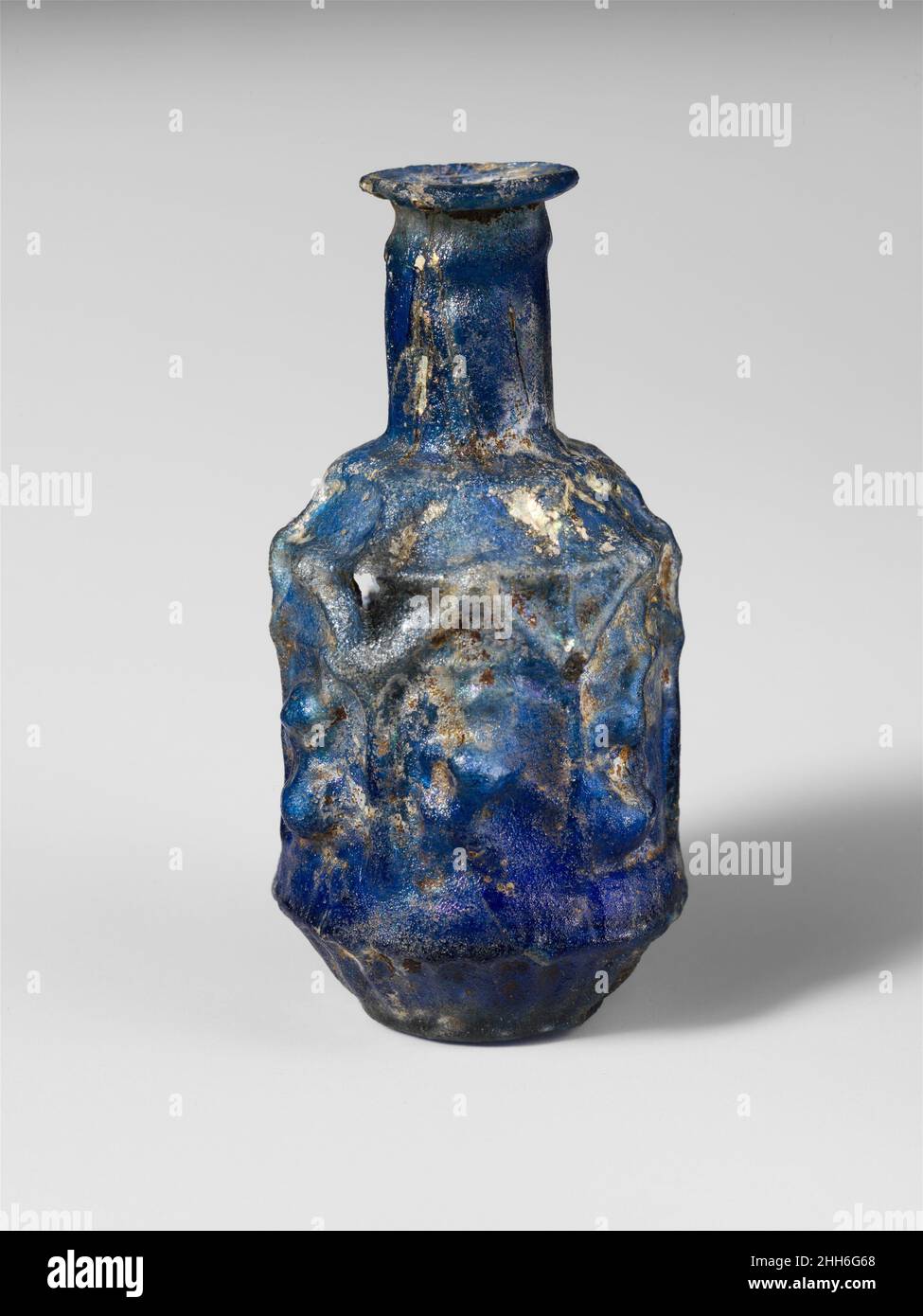 Glass hexagonal bottle ca. A.D. 25–50 Roman Translucent cobalt blueRim folded out, round, and in, then pressed into flaring mouth; cylindrical neck with concave band below rim; downward sloping shoulder; hexagonal body with vertical sides, then cup-shaped below; low base with rounded edge and flat but uneven bottom. Mold seams run from near top of neck, down sides, and meet off-center on bottom.Decoration in relief in three registers: on shoulder, six downturned rays or pointed petals; on body, six rectangular panels, each decorated at top with a triangular pediment, framed at sides with posts Stock Photo