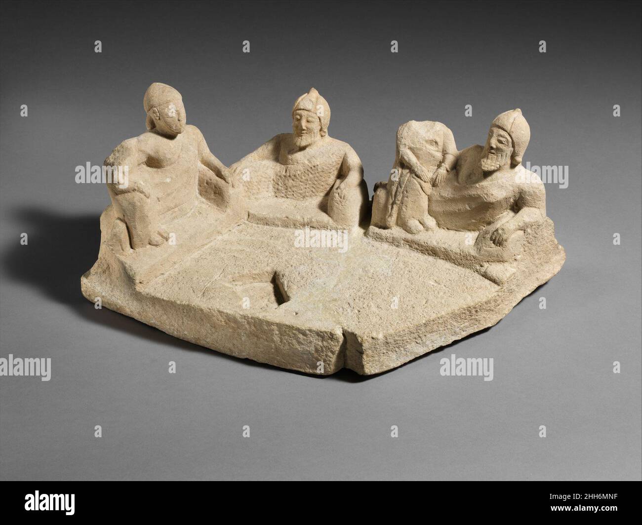 Limestone group: banquet end of the 6th century B.C. Cypriot Five figures are disposed around the edge of an irregular plinth. They face inward; a cutting may have held an altar or offering table. The banqueters recline on cushions, not on couches as in Greece. The central male figure is alone. On either side is a couple, a reclining male and a woman seated at his knees. The heads probably do not belong with the bodies. As with the terracotta group scenes, the Cypriot artist used all means at his disposal to render familiar subjects. The malleable clay and soft limestone facilitated his effort Stock Photo