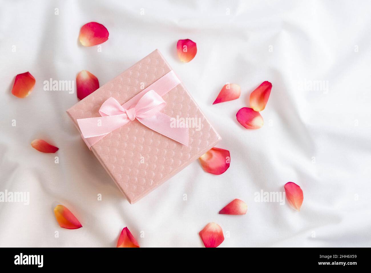 Top view of pink gift box with rose petals on white bed sheets. Romantic Valentines Day surprise and present Stock Photo