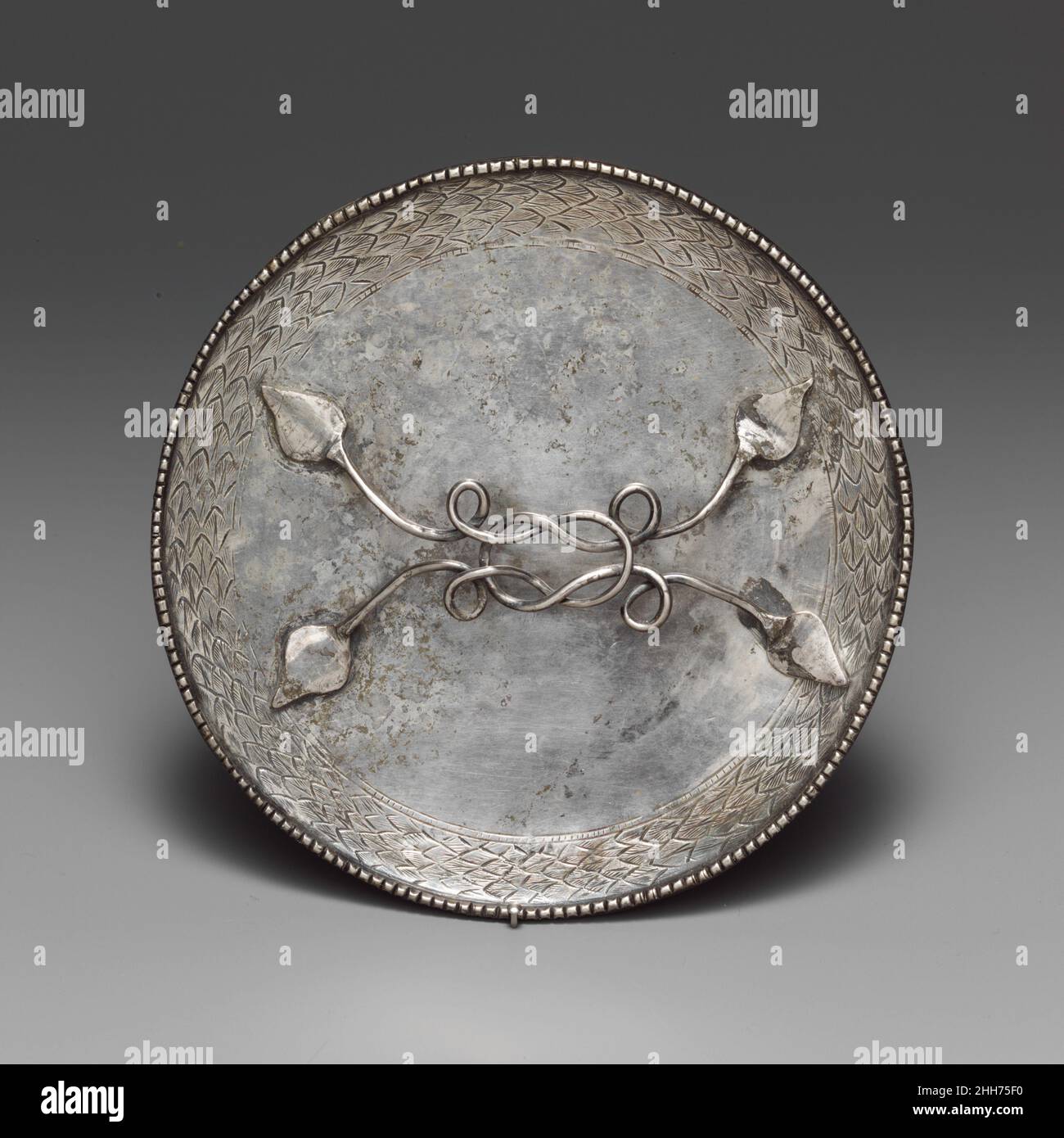 Silver mirror 4th century A.D. Roman The type of mirror with a horizontal handle originated in the Roman world during the first century B.C. It was subsequently adopted in various cultures of Asia and finally died out in the ninth to tenth century A.D. In this example, the classical origins are clear in the leaf-shaped attachments of the handle, the Herakles knot, and the wreath around the circumference of the disk.. Silver mirror. Roman. 4th century A.D.. Silver. Imperial. Gold and Silver Stock Photo