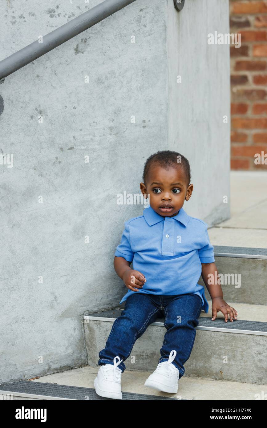 A cute one year old toddler almost preschool age African-American boy with big eyes sitting on city steps Stock Photo