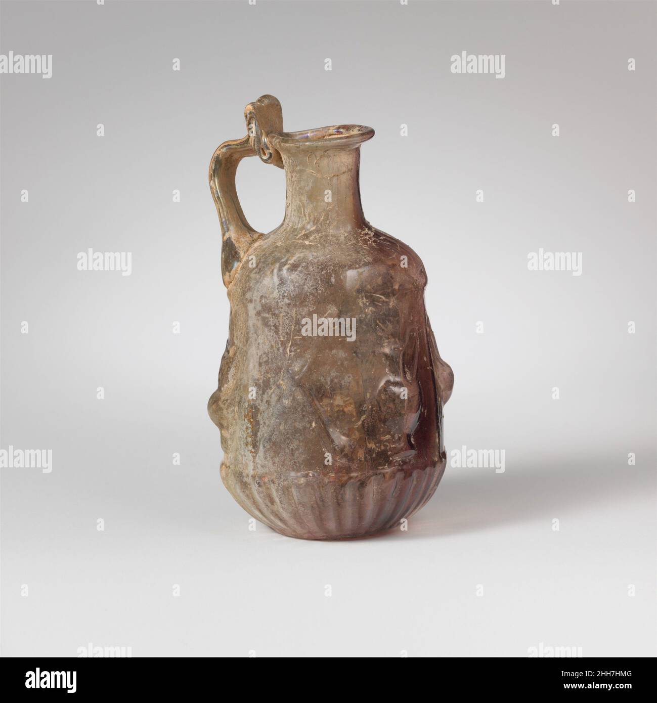Glass hexagonal jug 1st century A.D. Roman Colorless with translucent purple streak from rim to bottom, with colorless handle.Tubular rim, folded out, round, and into mouth; concave cylindrical neck; sloping, rounded shoulder; straight-sided hexagonal body, expanding downward; convex undercurve; flat circular bottom; strap handle attached in two large ribs to shoulder and top edge of body above division between panels 5 and 6 (below), drawn up and slightly out, then curved in, and pressed on to top of neck and underside of rim, with projecting flattened thumb rest above.On shoulder, frieze of Stock Photo