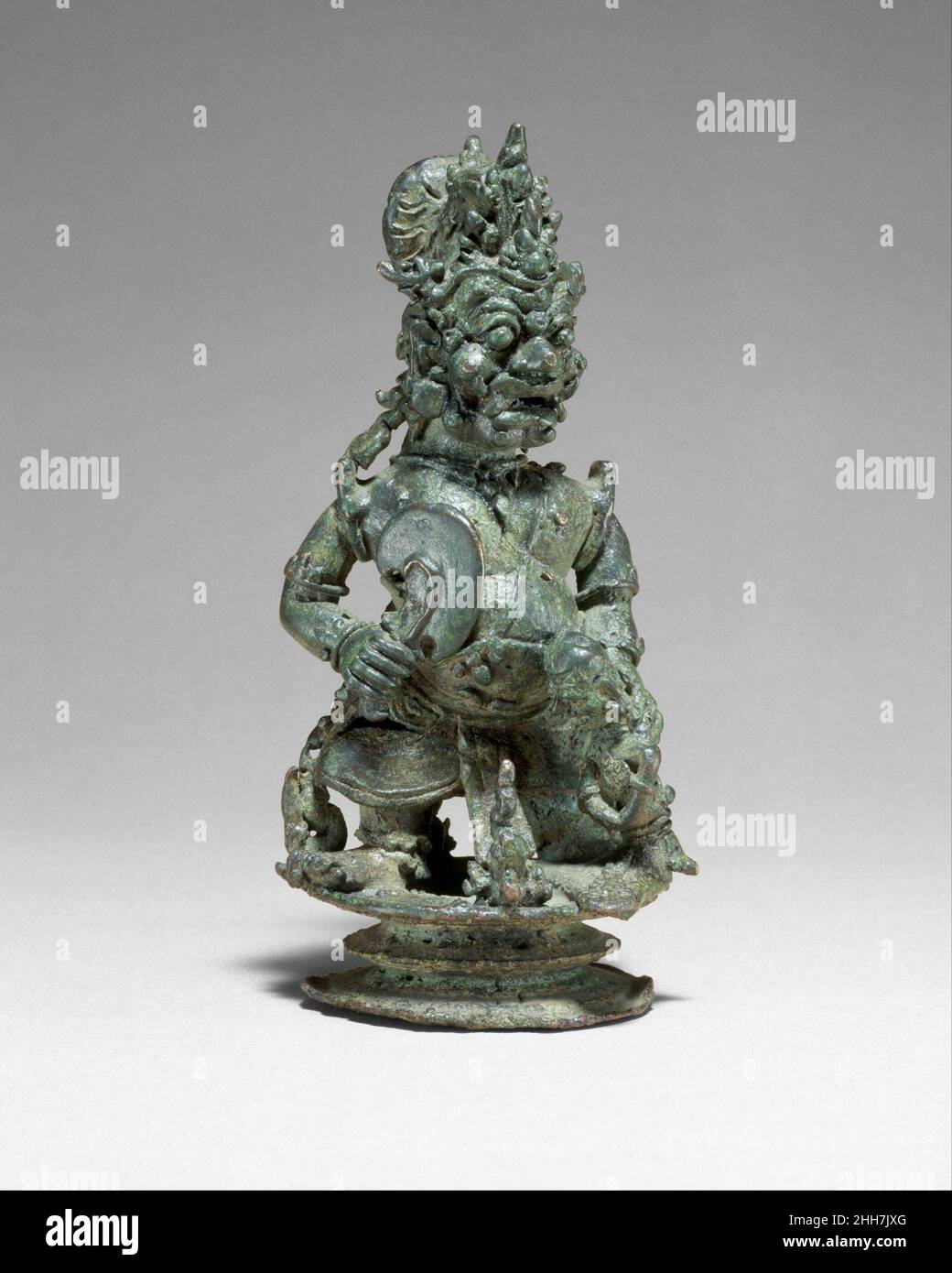 Top of a Bell in the Form of a Demon King or Guardian ca. second half of the 12th–early 13th century Indonesia (Java) This finial from a hanging bell takes the form of an unusually lively and finely modeled rakshasa—a demon king or guardian. He is depicted as a short, potbellied grimacing creature with fangs and large bulbous eyes and a serpent emerging from each armpit. A curved broad chopper is held in his right hand, and his left is placed behind the neck of a hapless victim with bound hands who is seated in front of him. The hair is pulled back and arranged in a loop to allow for the attac Stock Photo