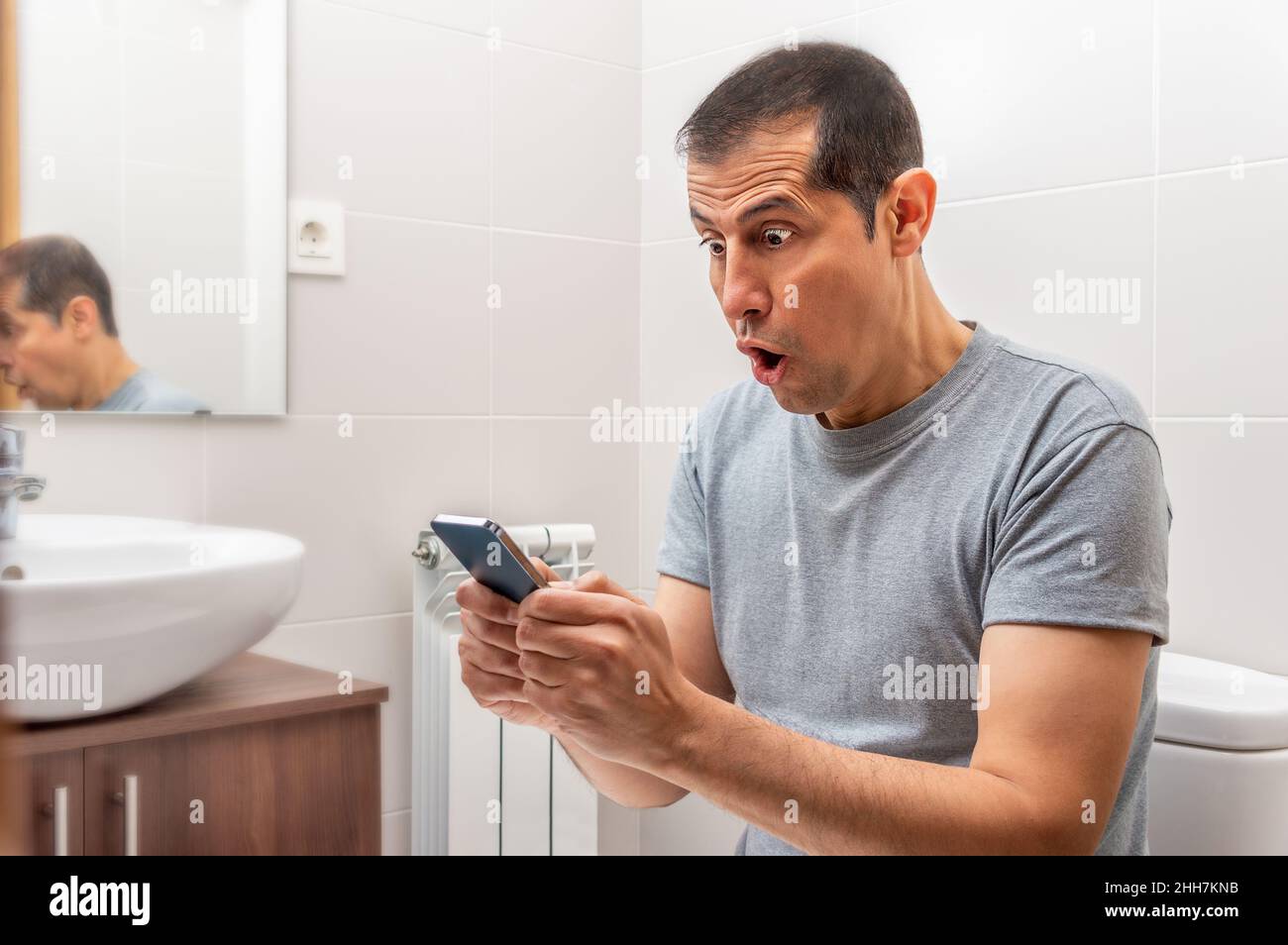 Latin man receiving surprising news on line in a smart phone at the bathroom. Stock Photo