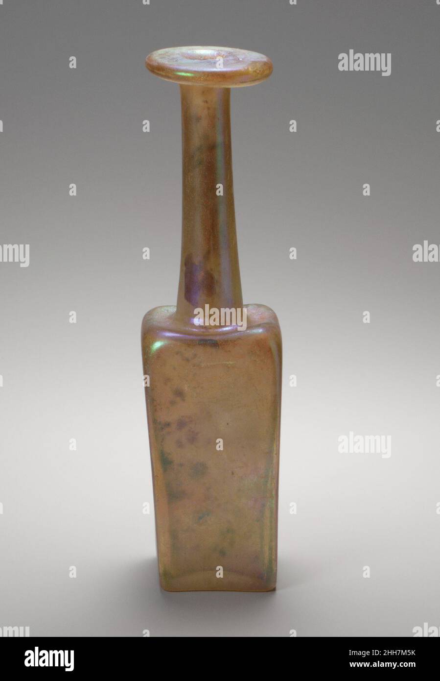 Glass 'Mercury' bottle 3rd century A.D. Roman Colorless with pale yellow green tinge.Broad rim, folded out, down, round and in, and flattened on top surface; thick-walled cylindrical neck, expanding downwards; pushed-in horizontal shoulder with raised rounded corners; square, thick-walled body, with flat sides; concave bottom.In relief on bottom, standing peacock, facing right, with crest above head and tail feathers displayed behind in a stylized fashion with two circular rows of dots.Intact; dulling, pitting, and brilliant iridescence covering almost all of surfaces.Bottles of this type are Stock Photo
