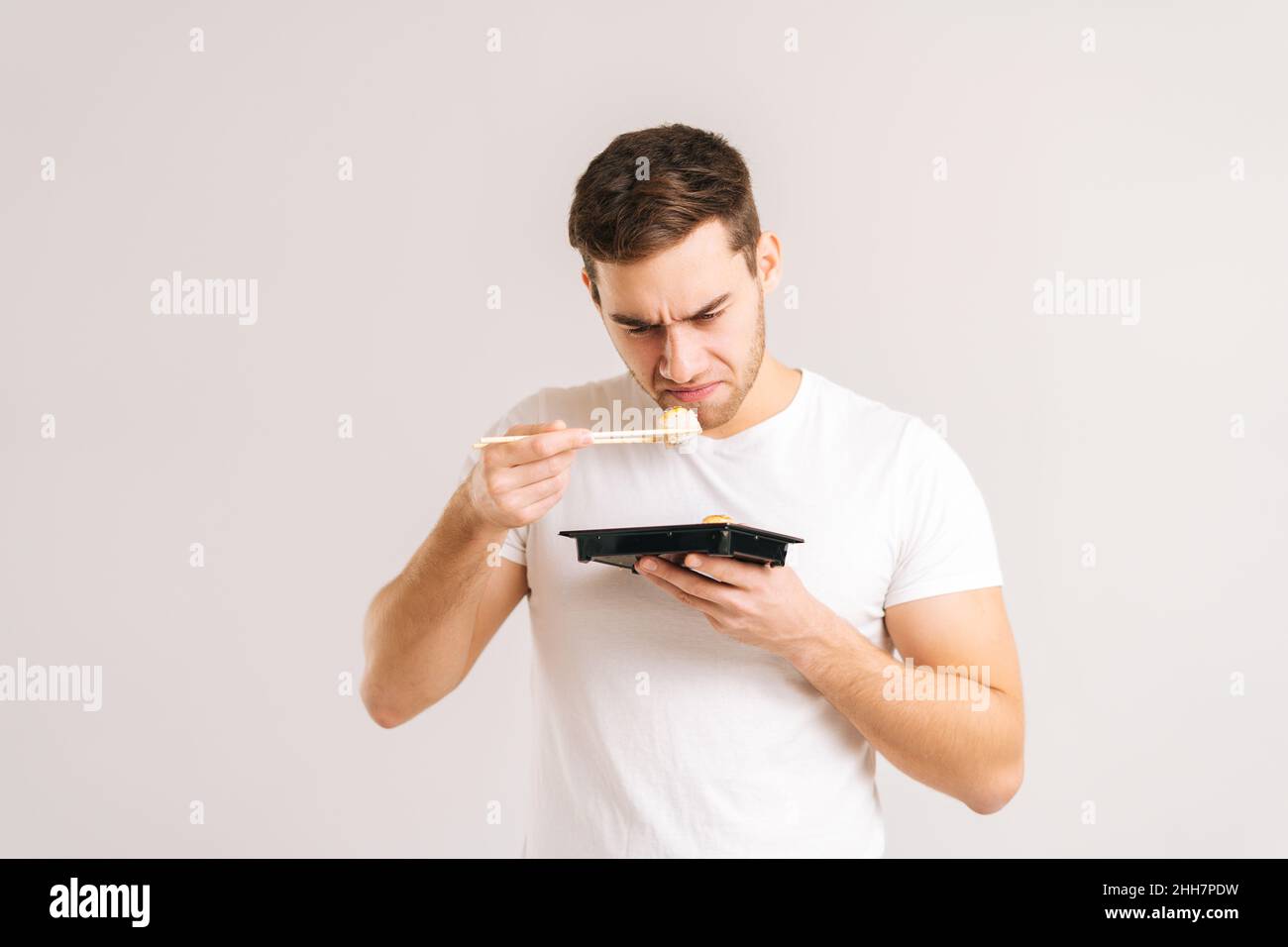 Portrait of disgusted young man holding sushi with chopsticks and sniffs with look of revulsion on face on white isolated background. Stock Photo