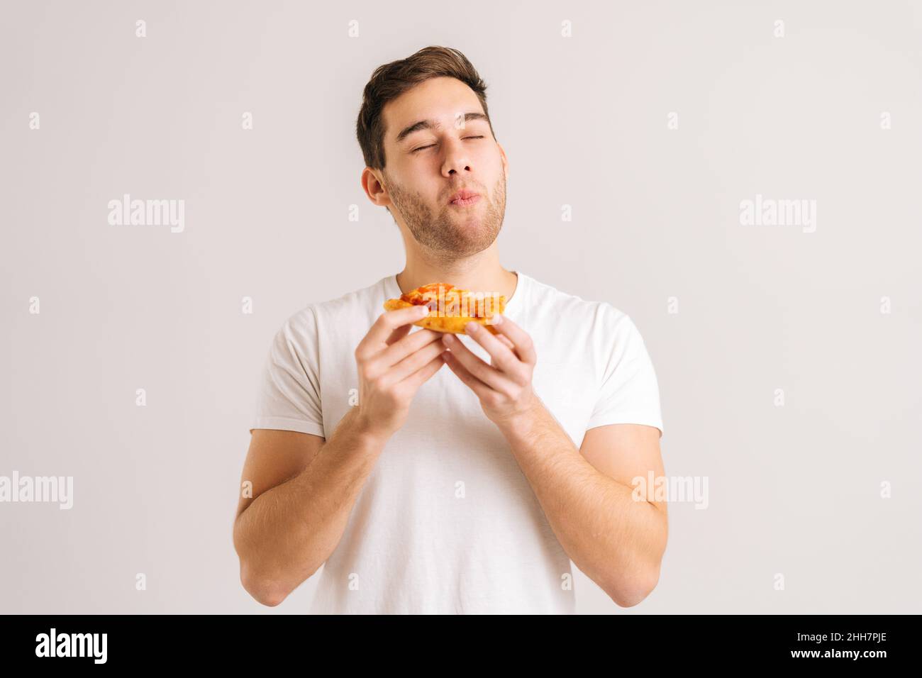 Portrait of handome young man with enjoying eating delicious slice of pizza, with closed eyes from pleasure on white isolated background. Stock Photo