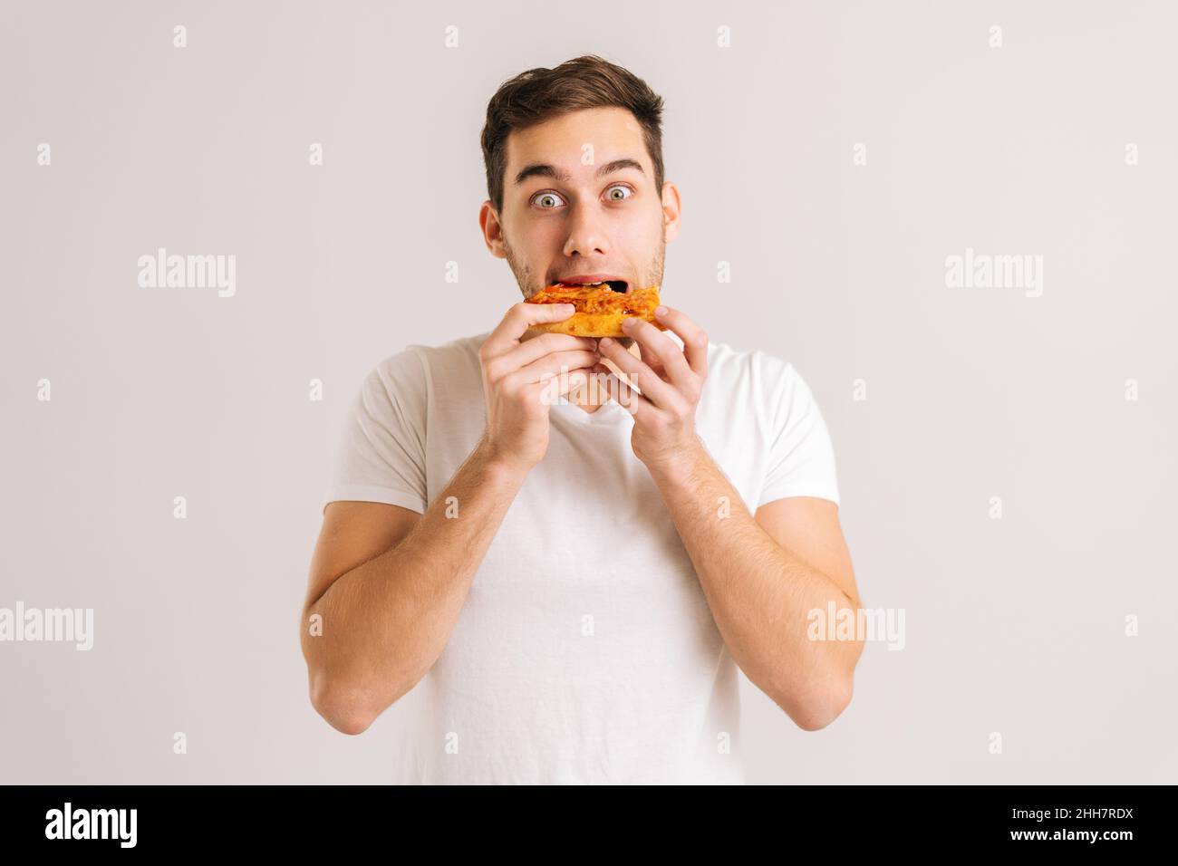 Portrait of exctited young man bitting delicious slice of pizza looking at camera on white isolated background. Stock Photo