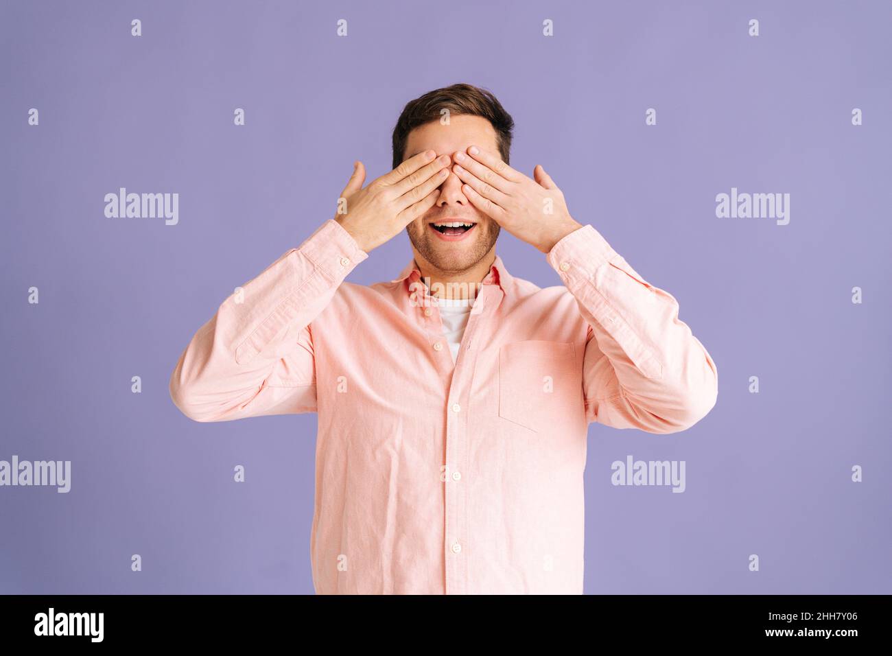 Portrait of handsome young man covering eyes by hands and smiling over ...
