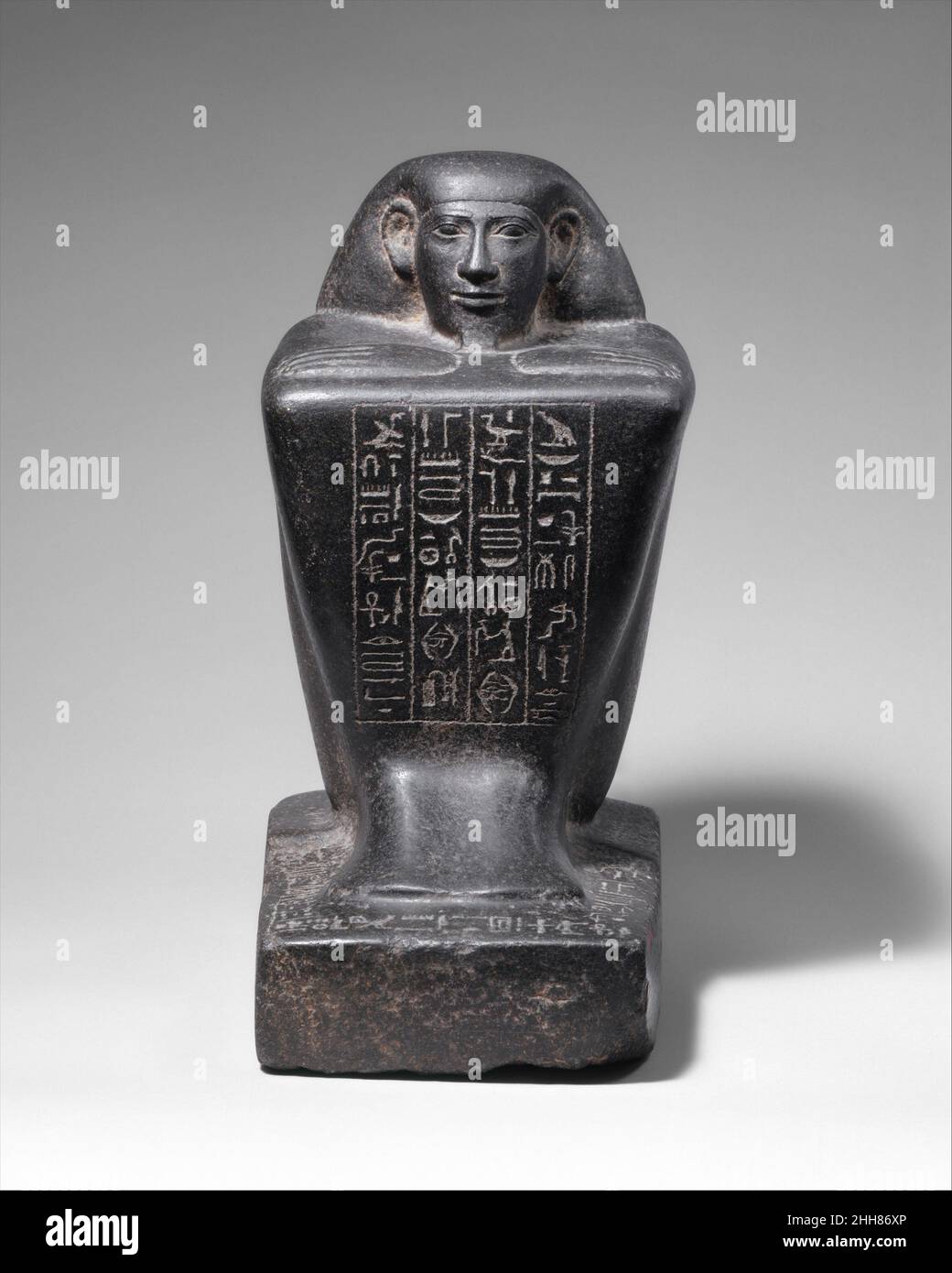 Block Statue of a Prophet of Montu and Scribe Djedkhonsuefankh, son of Khonsumes and Taat 690–610 B.C. Late Period, Kushite–Saite In the late 25th dynasty and early 26th Dynasty the austere beauty of the block statue form, here particularly enhanced by the hard gleam of dark surfaces, was greatly appreciated. The form puts a strong emphasis on the face and complements the often stern countenances favored at the time.The front of the statue gives the names, titles and parentage of Djedkhonsuiuefankh, son of Khonsumes and Taat. Djedkhonsuiuefankh can be linked to a multigeneration illustrious fa Stock Photo