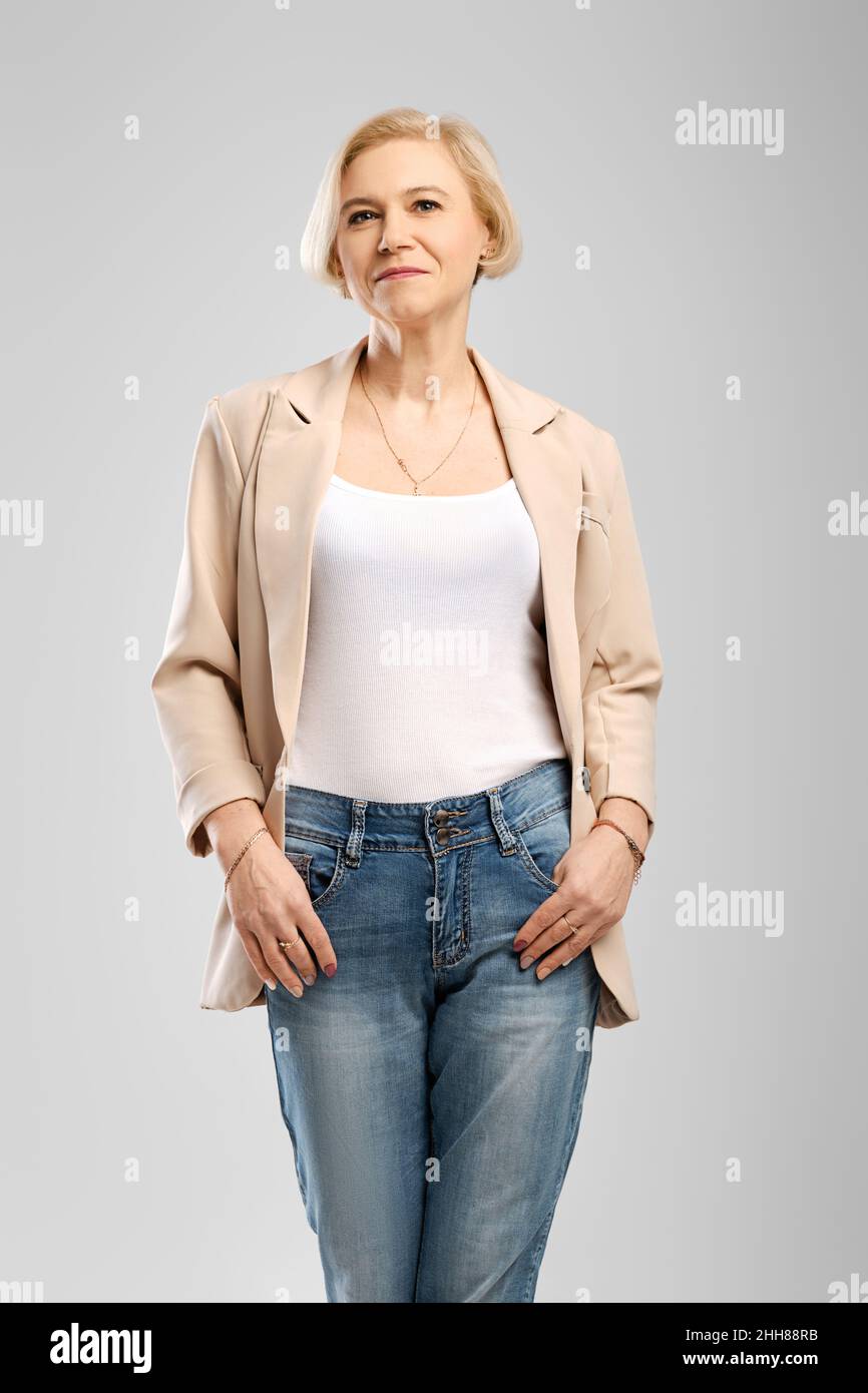 Stylish confident senior female standing with arms in the pockets looking at camera at gray background Stock Photo