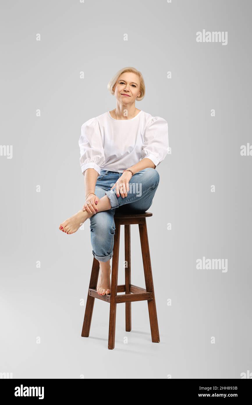 Barefoot senior woman in jeans and white blouse sitting on tall chair in studio throws one leg Stock Photo