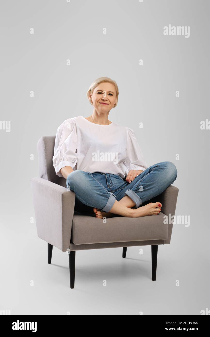 Barefoot senior woman sitting in an armchair crossing legs in studio Stock Photo