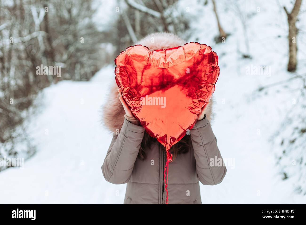 Girl in fur jacket holding red helium heart shaped balloon in front of her face Stock Photo