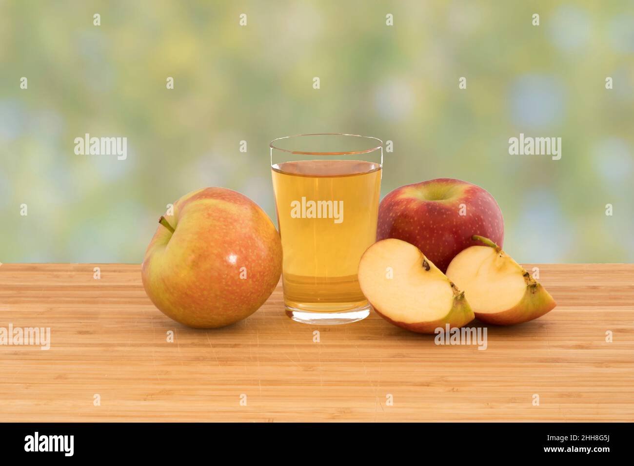 A glass of apple juice with apples and apple slices on a wooden cutting board isolated on a green bokeh background Stock Photo