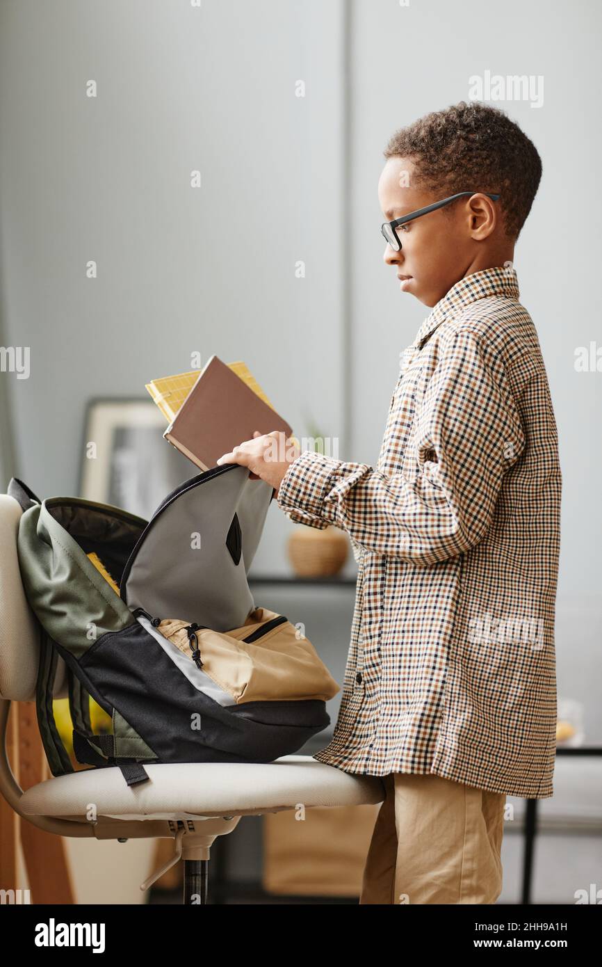 Vertical side view portrait of young African-American schoolboy packing backpack at home Stock Photo