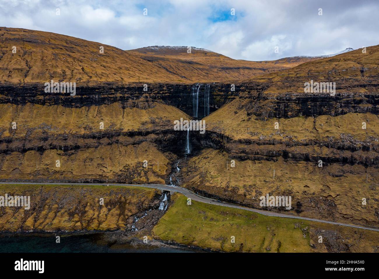 The waterfall finds its path through the stepped rocks, covered in vegetation, and goes under the road. Stock Photo