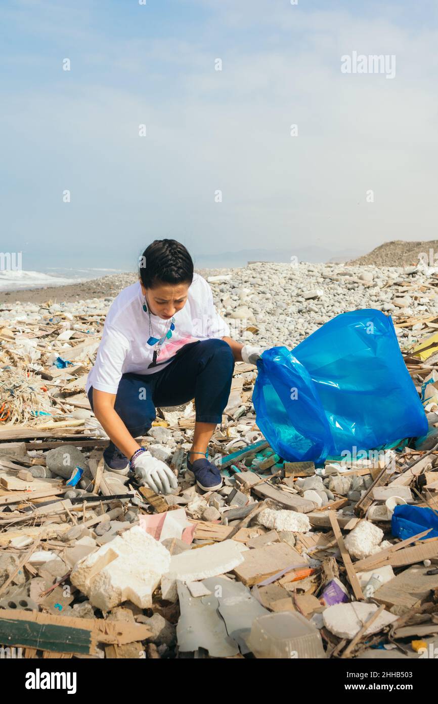 Vertical image of a latin woman volunteer picking up trash on a polluted beach. Stock Photo