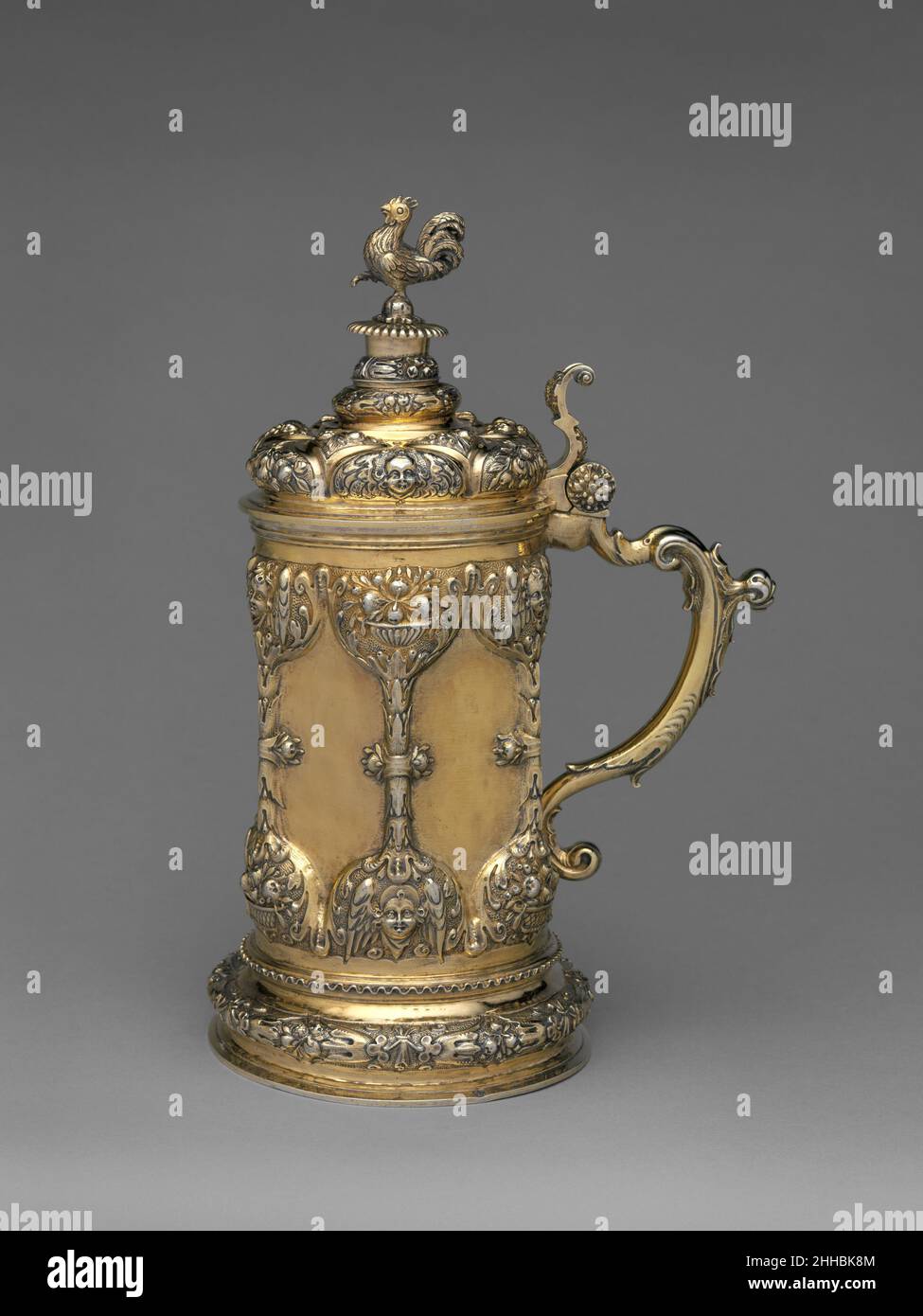 Tankard late 17th century Georgius Olescher Sr. It is unclear if this tankard was made for private use or if it was always intended to serve as a communion vessel, which was its function after it arrived in England. The overall ornamental symbolism is connected to vanity. Fruit and foliage represent life, and stylized hourglass motifs divide the main body into six plain panels (for this vanity tradition see, Wolfram Koeppe. Die Lemmers-Danforth Sammling Wetzlar. Heidelberg, 1992, pp. 464-465, cat. GO 8). The cast finial plinth supports a cockerel standing on one foot, a pose adopted by the bir Stock Photo